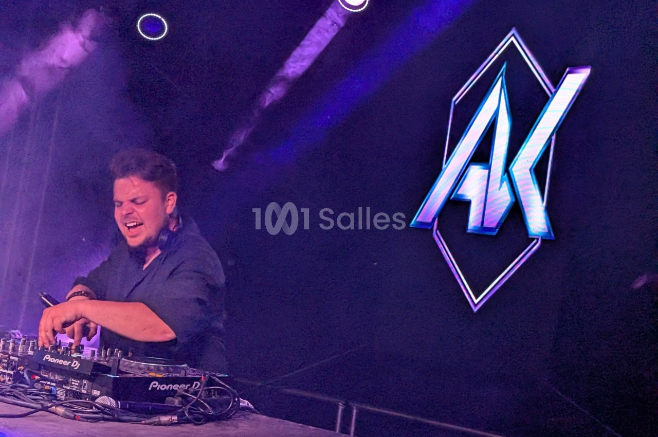 Un DJ mixe sur scène, entouré de lumières violettes, avec un logo lumineux ’AK’ en arrière-plan.