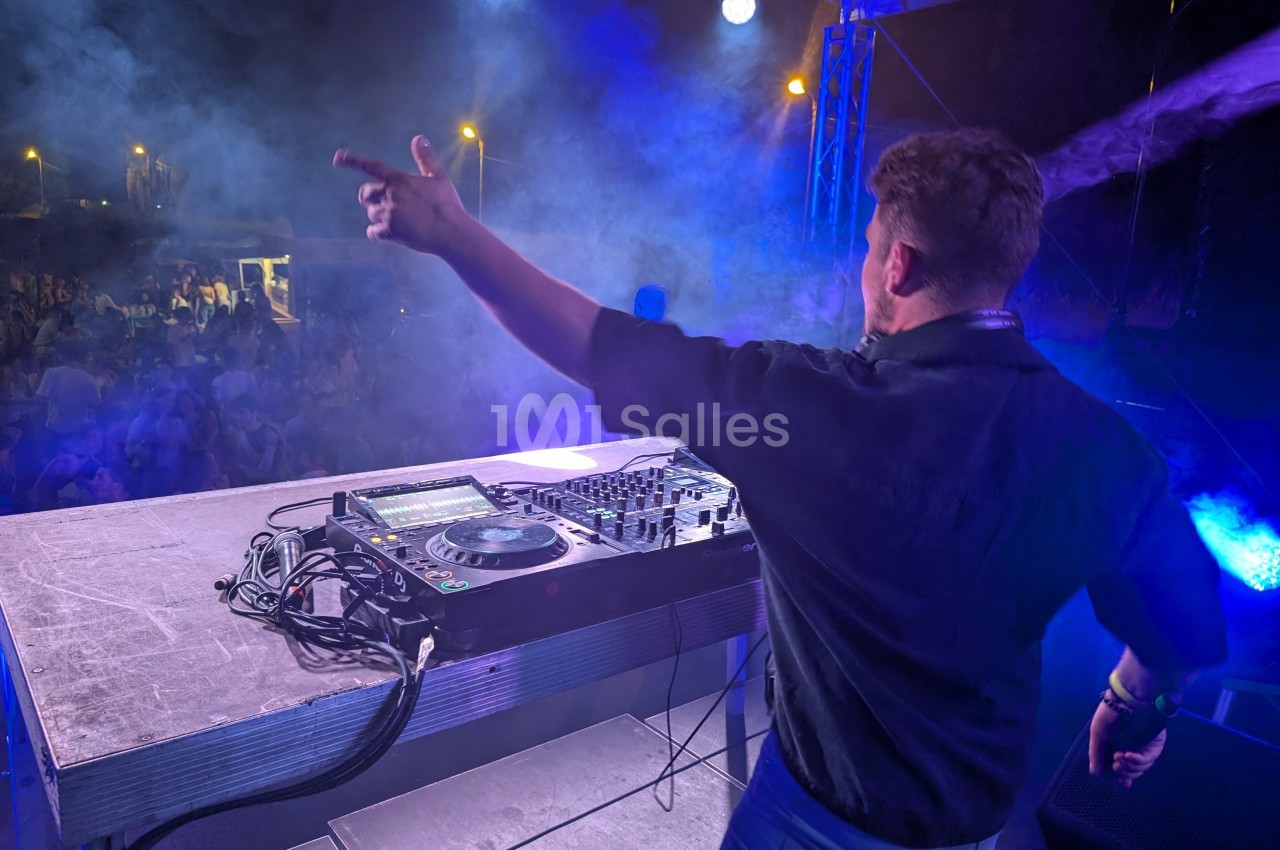 Un DJ anime une foule depuis une scène éclairée, entourée de fumée et d'effets lumineux.