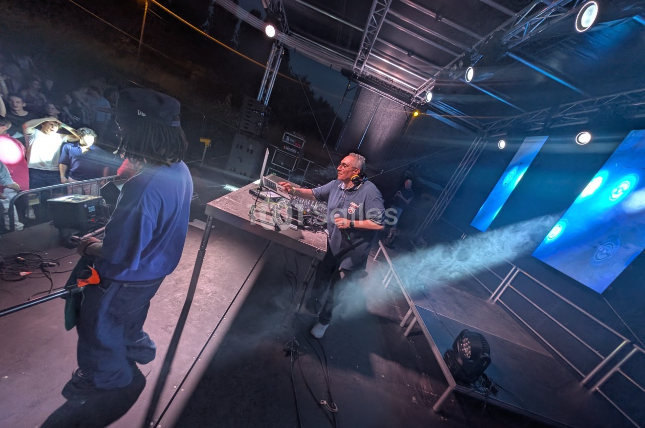 Un DJ mixe sur scène sous des éclairages colorés, avec un public en arrière-plan.