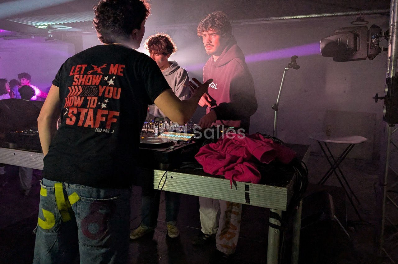 Un homme en t-shirt noir échange avec deux autres personnes près d'une table de mixage dans une salle éclairée par des…