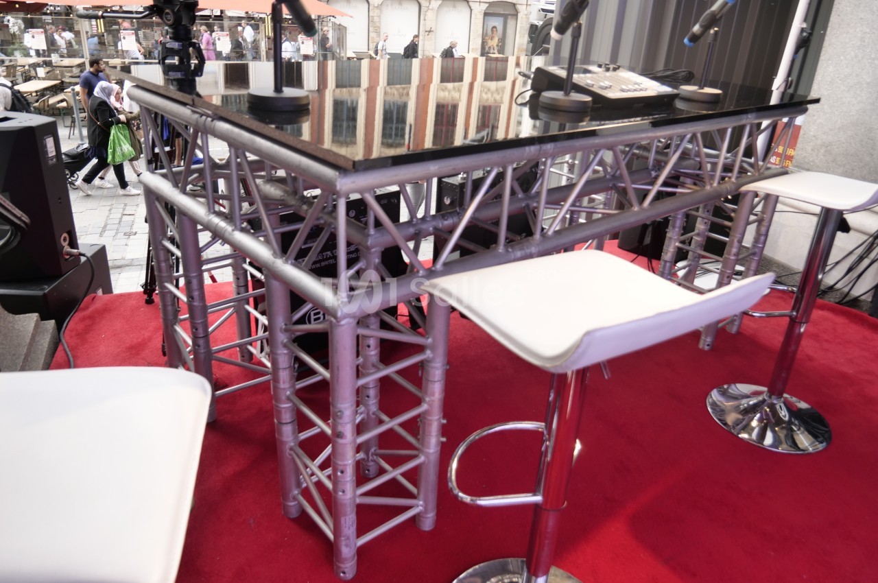 Table haute en verre sur une structure métallique, entourée de tabourets blancs, installée sur une scène avec des micros.