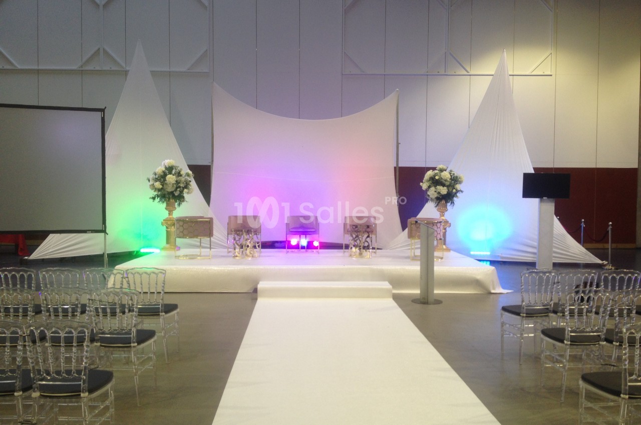 Scène décorée pour un événement avec chaises transparentes, fleurs, éclairages colorés et fond en tissu blanc.