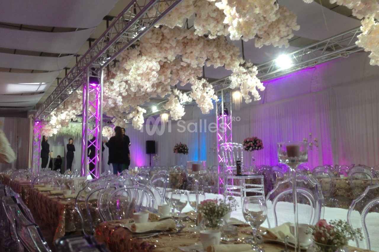 Salle de réception décorée avec des tables dressées, des chaises transparentes et des fleurs suspendues blanches.