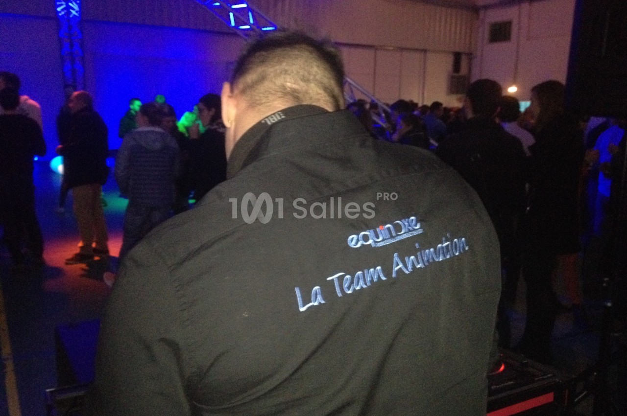 Un animateur de dos portant une chemise avec un logo, dans une salle éclairée par des lumières colorées.