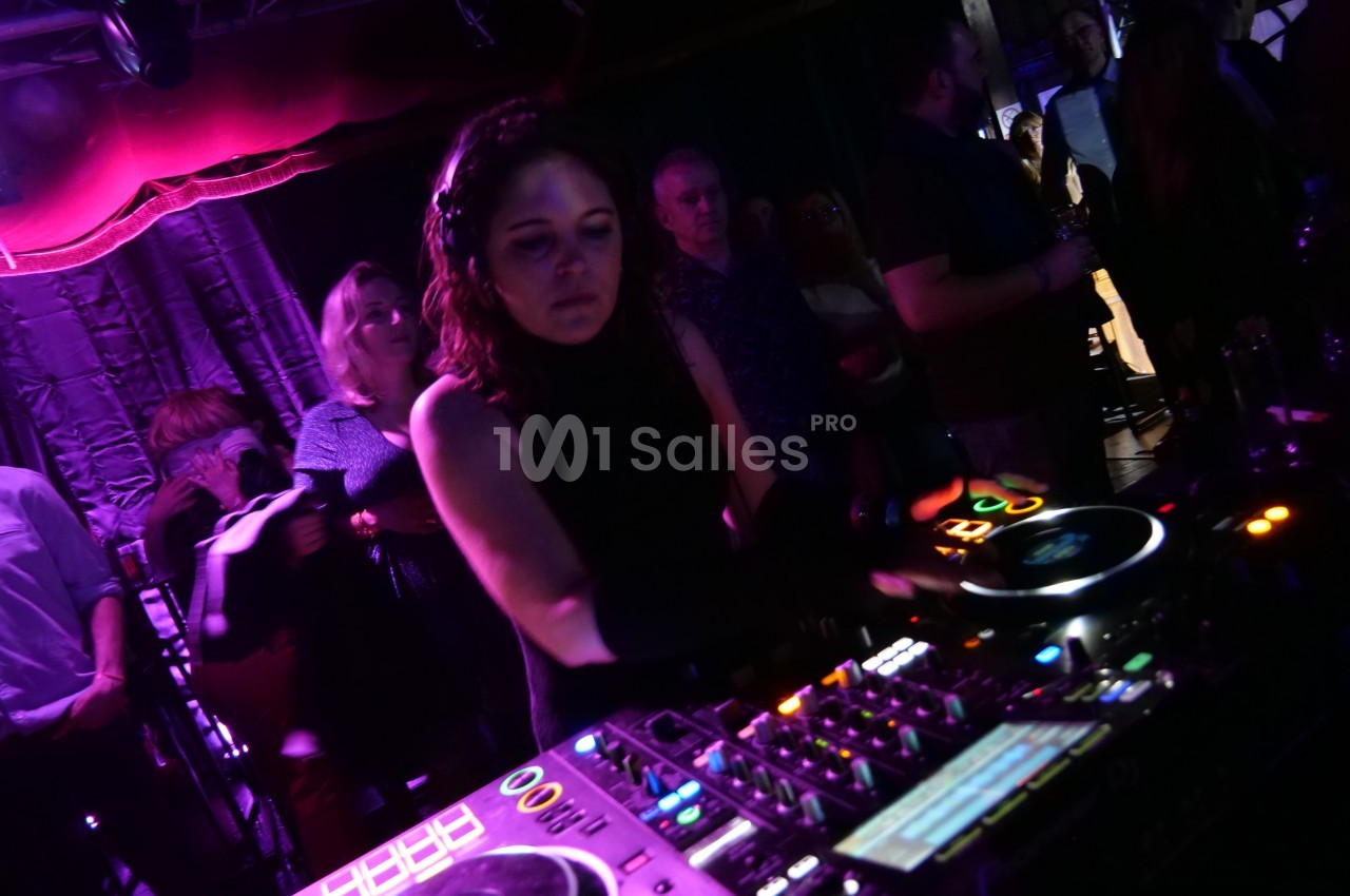 Une DJ mixe sur une console dans une ambiance sombre avec des lumières violettes, entourée de spectateurs.