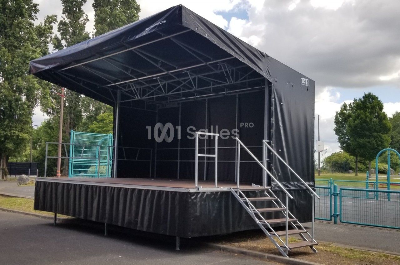 Scène mobile extérieure avec structure métallique et bâche noire, installée dans un espace ouvert près d'une aire de jeux.