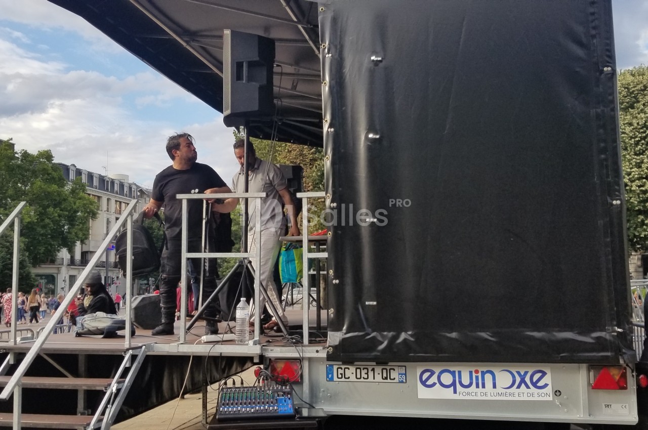 Deux hommes sur une scène mobile en plein air, avec du matériel audio, entourés de spectateurs et d'arbres.