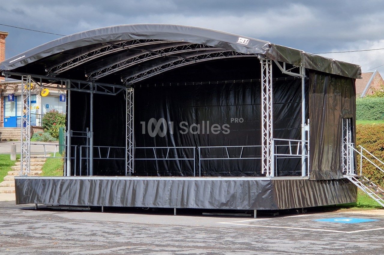 Scène mobile couverte avec structure métallique, installée sur un parking sous un ciel nuageux.