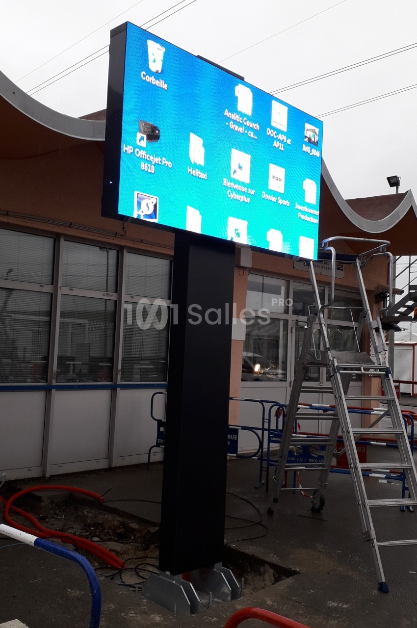 Écran numérique monté sur un poteau devant un bâtiment, affichant une interface d'ordinateur avec des icônes de dossiers.