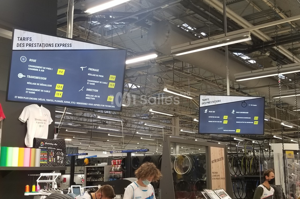 Écrans affichant les tarifs des prestations dans un atelier de réparation, avec employés et clients en arrière-plan.