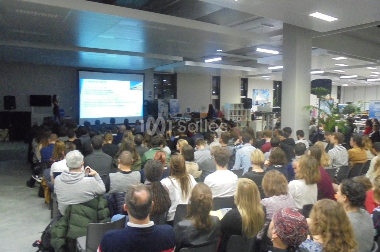 Salle comble avec des participants assis, assistant à une présentation projetée sur un écran dans un espace moderne.