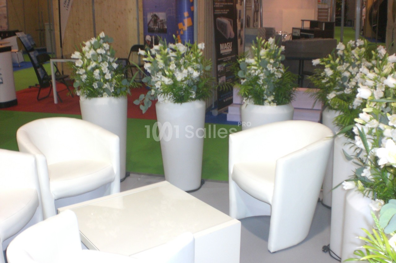 Chaises blanches et tables basses entourées de grands vases avec des arrangements floraux dans un espace d'exposition.