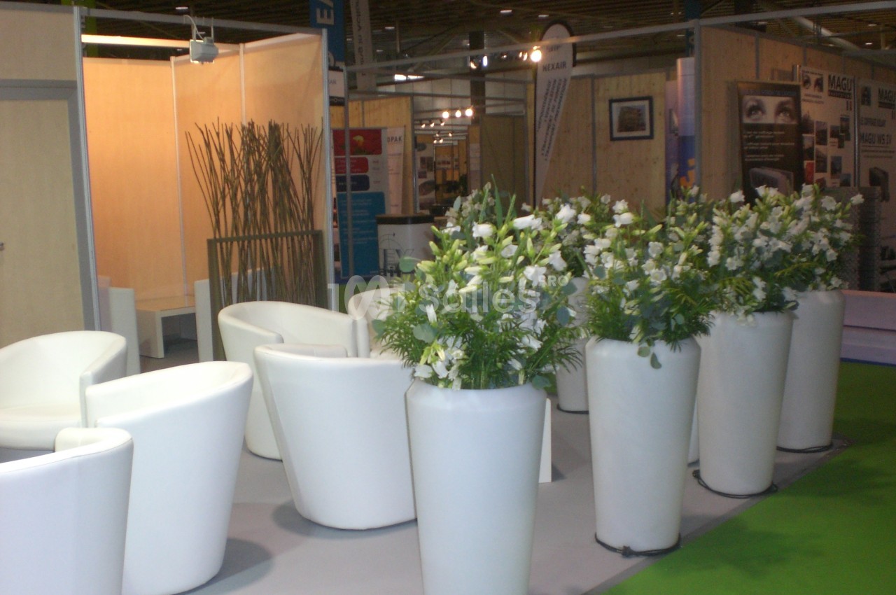 Des fauteuils blancs entourent des plantes en pots dans un espace d'exposition au sol gris et vert.