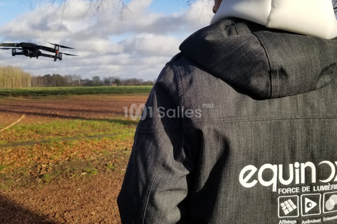 Une personne pilote un drone en plein vol au-dessus d'un champ, par une journée partiellement nuageuse.