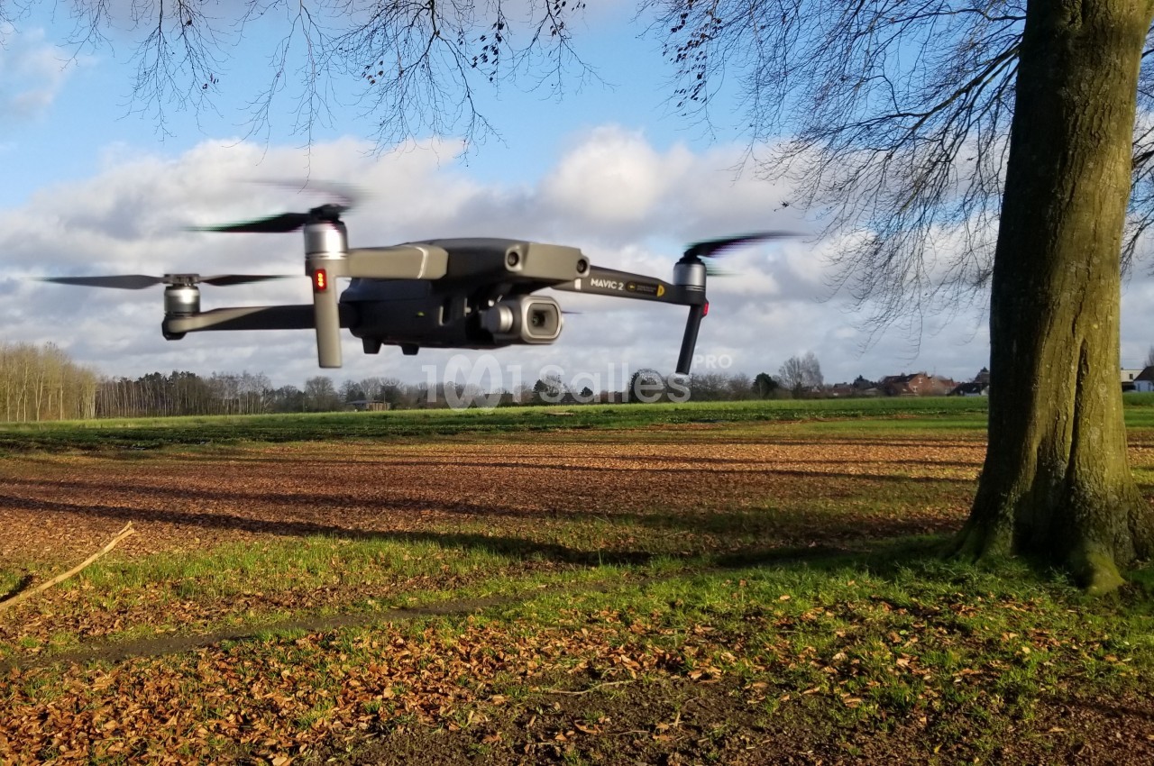 Un drone gris en vol stationnaire au-dessus d'un champ, près d'un arbre, sous un ciel partiellement nuageux.