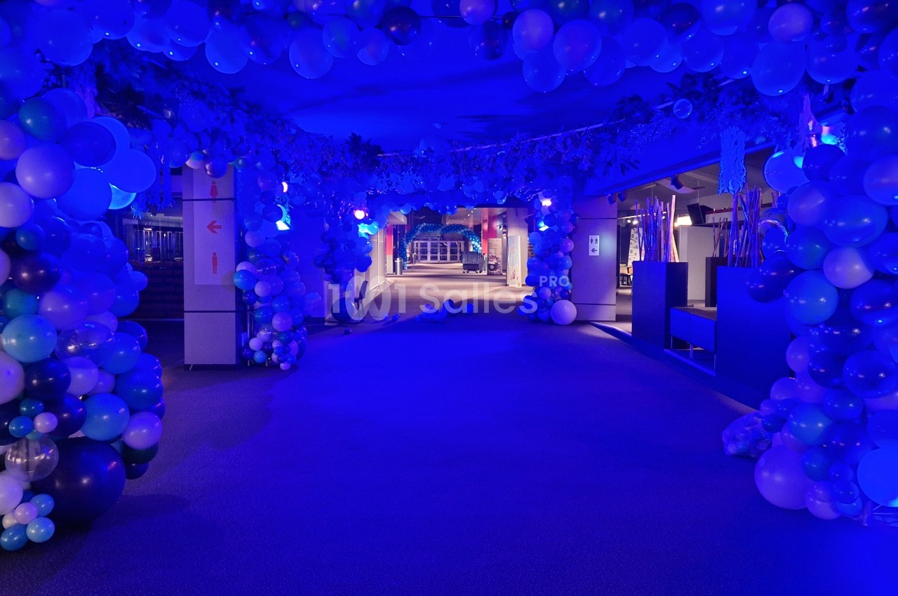 Couloir décoré de ballons bleus et blancs éclairé par des lumières bleues, créant une ambiance festive.