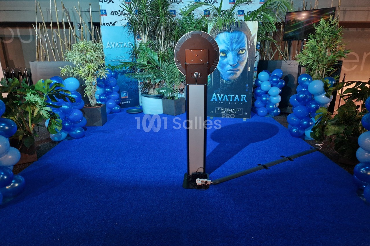 Stand photo sur tapis bleu entouré de plantes, ballons bleus et affiche promotionnelle d'un film en arrière-plan.