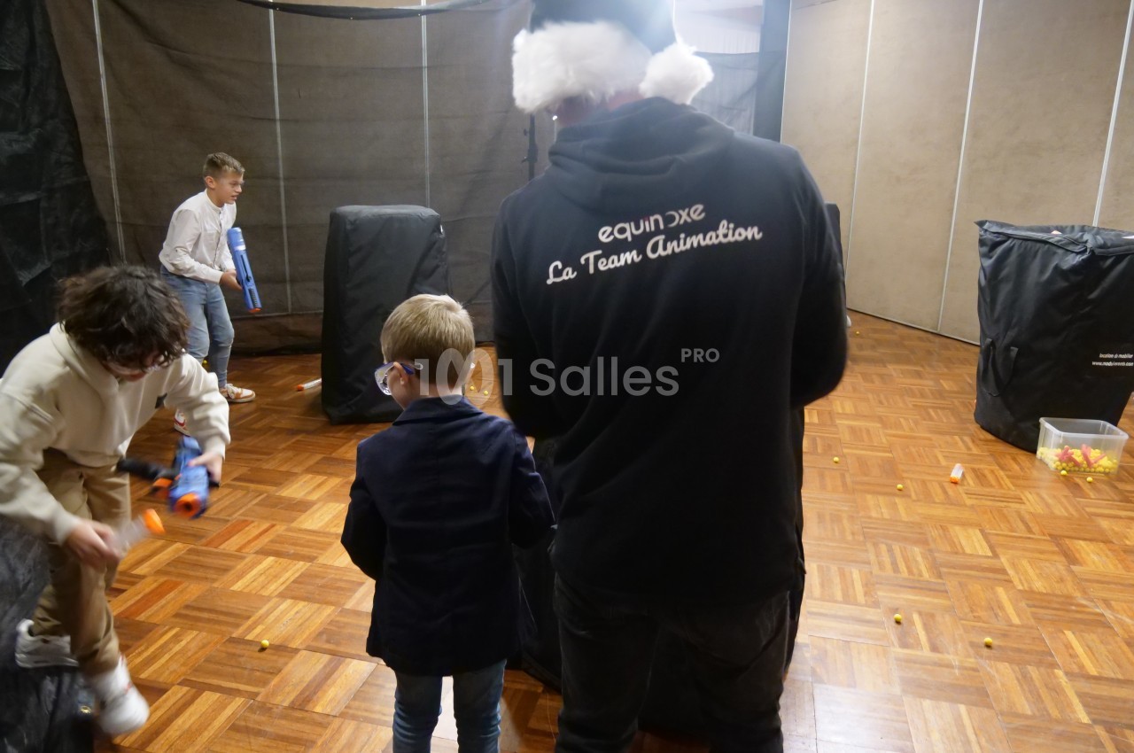 Des enfants jouent avec des pistolets en mousse dans une salle aménagée avec des obstacles et un animateur présent.