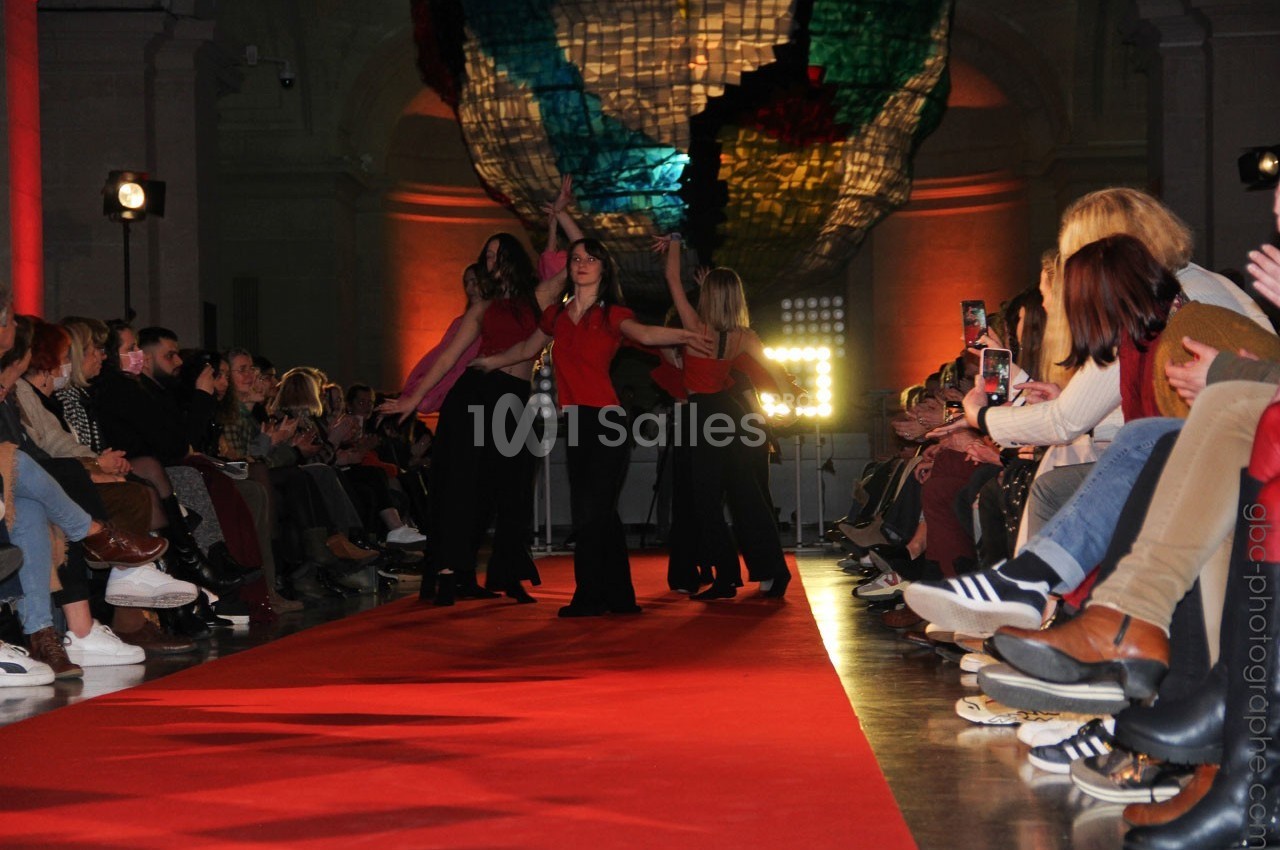 Des danseuses en tenue rouge et noire exécutent une chorégraphie sur un podium entouré de spectateurs.
