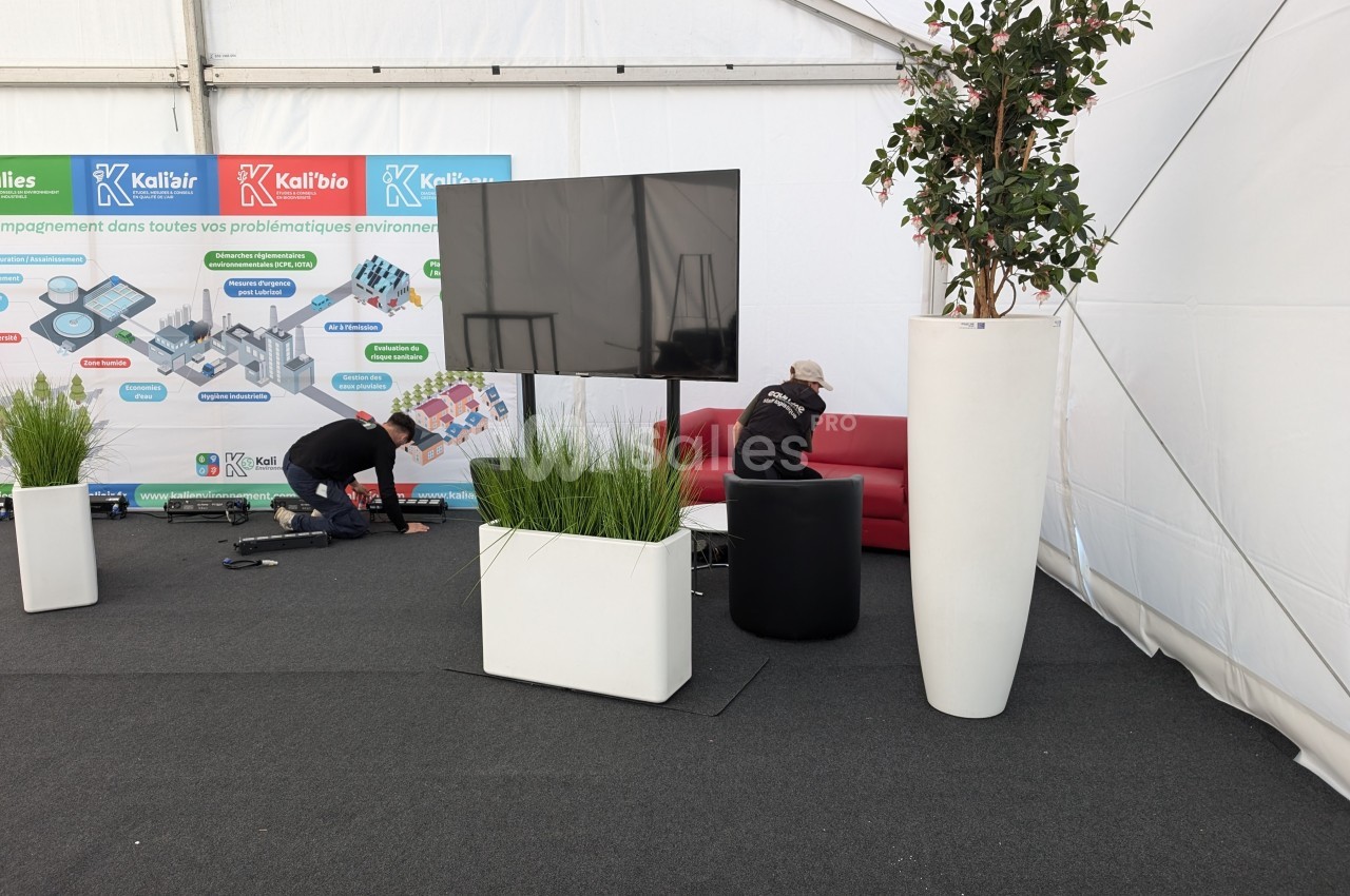 Un stand sous tente avec un écran, des plantes en pots, un canapé rouge et deux personnes installant du matériel.