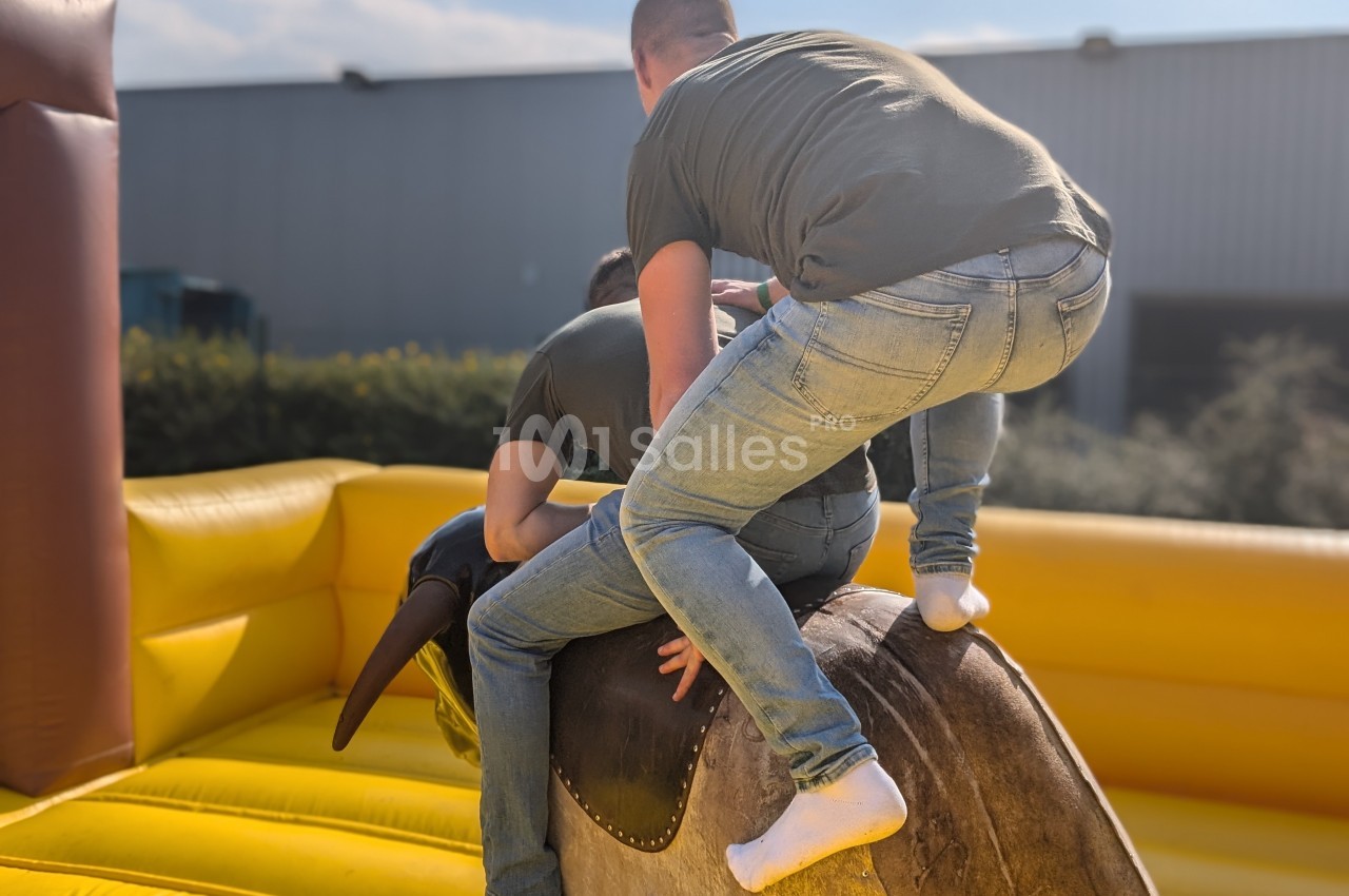 Deux personnes en jeans tentent de monter sur un taureau mécanique dans une structure gonflable en extérieur.