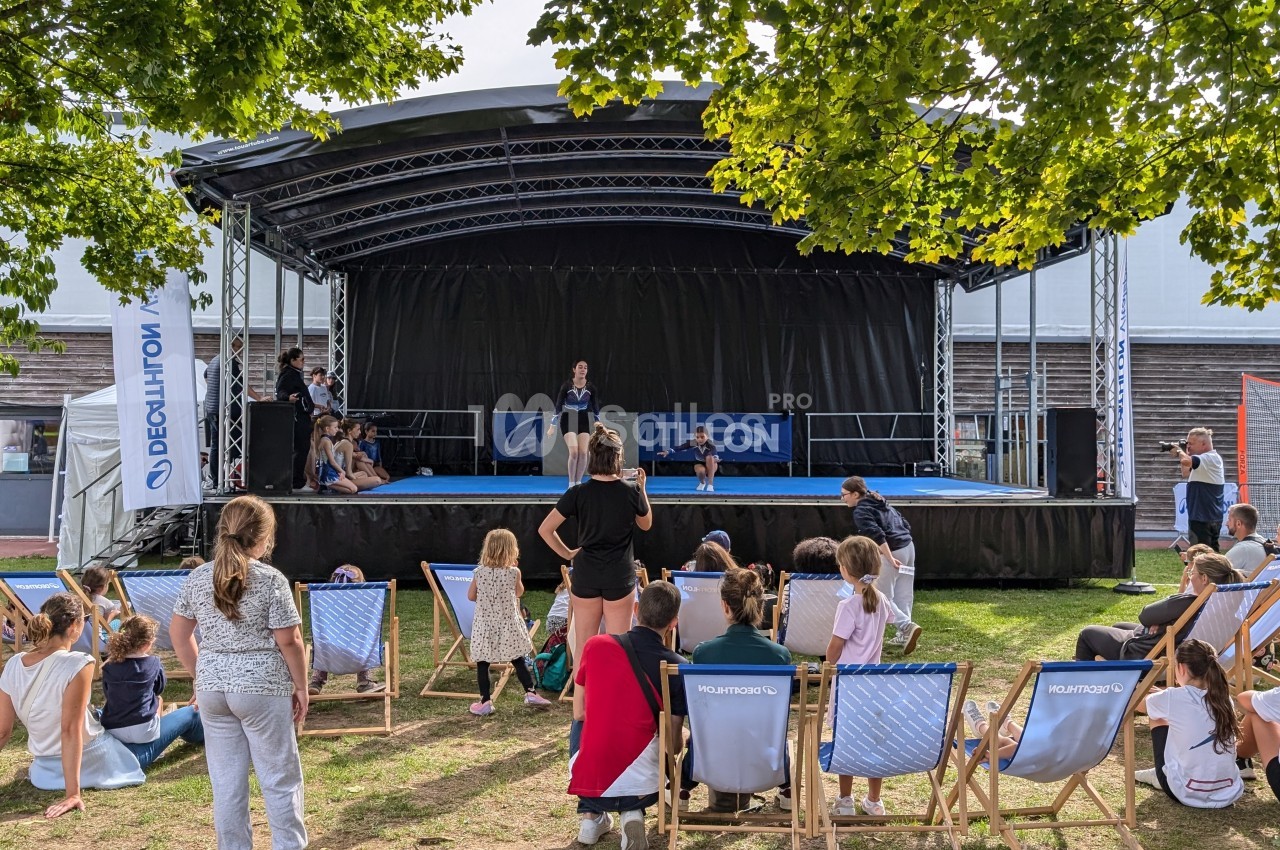 Des spectateurs assis devant une scène en plein air où se déroule une présentation, entourée d'arbres et d'installations.