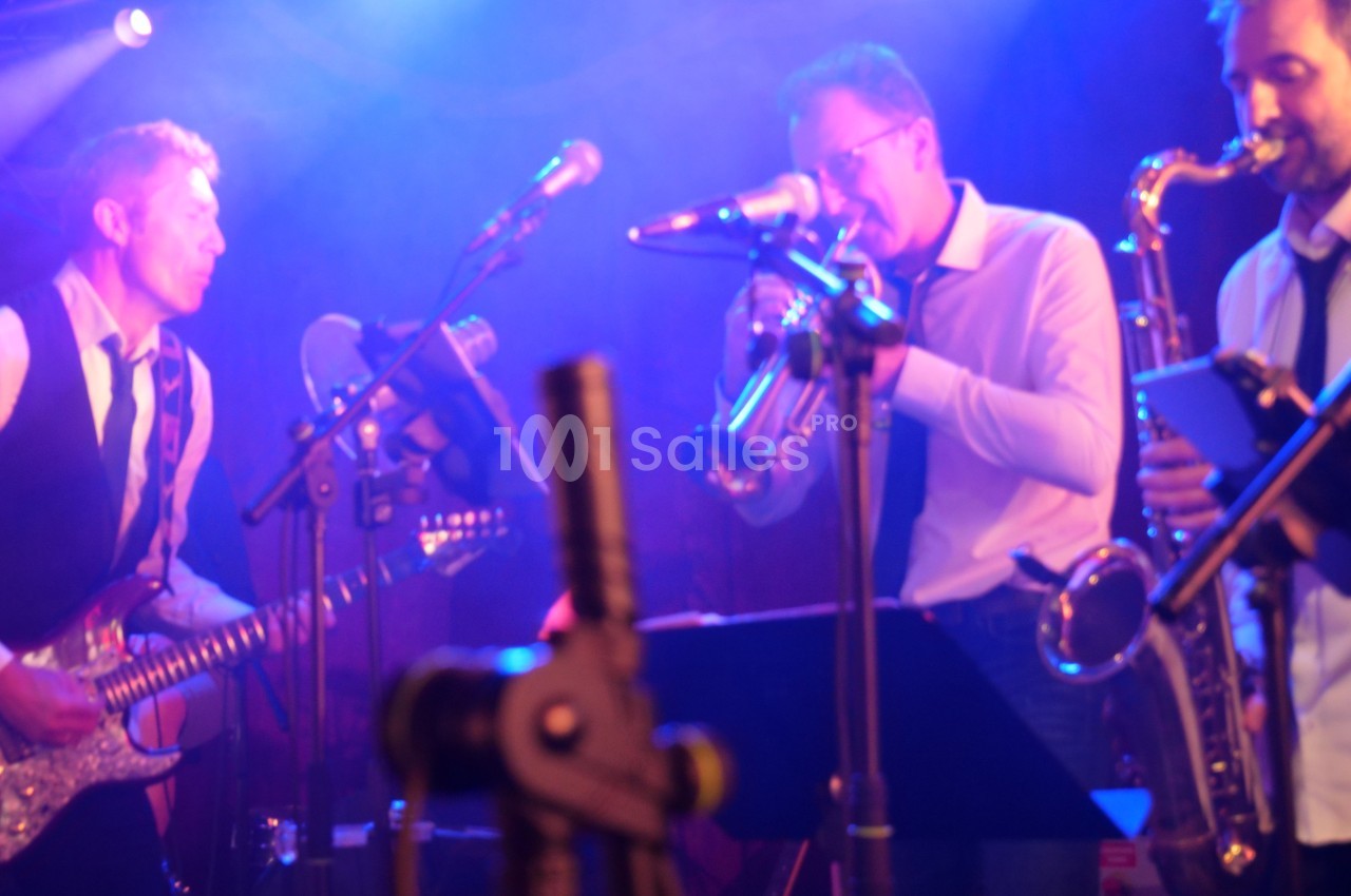 Musiciens jouant de la guitare, de la trompette et du saxophone sur une scène éclairée par des lumières colorées.