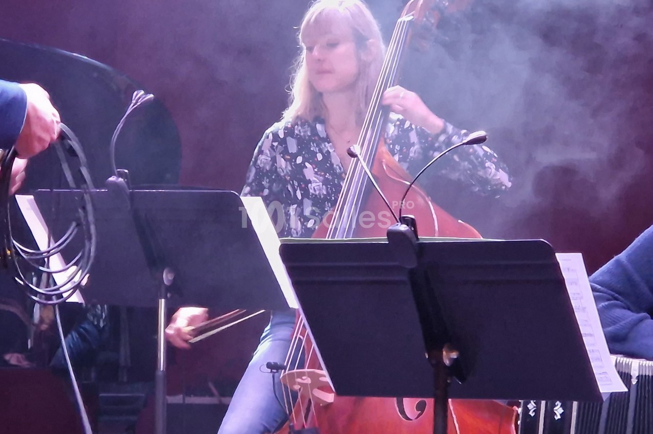 Une musicienne joue de la contrebasse sur scène, entourée de partitions et d'un léger brouillard lumineux.