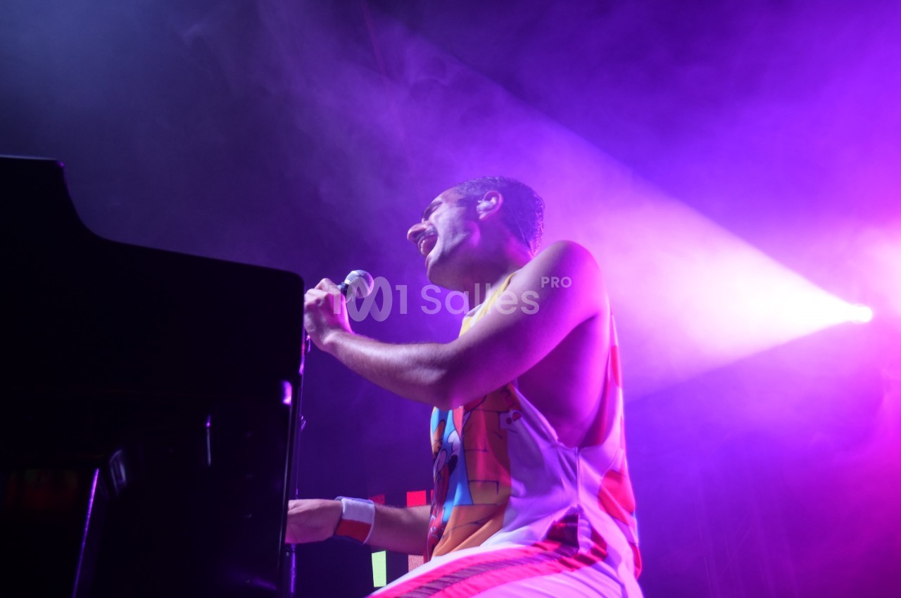 Un chanteur joue du piano sur scène sous des lumières violettes et un projecteur lumineux.