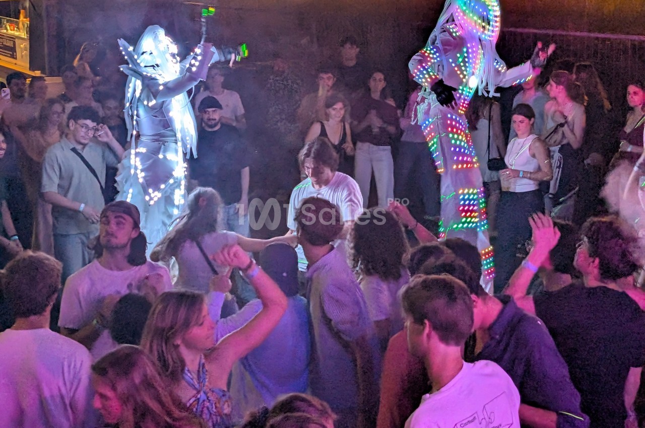 Des artistes en costumes lumineux dansent sur des échasses au milieu d'une foule lors d'une soirée festive.