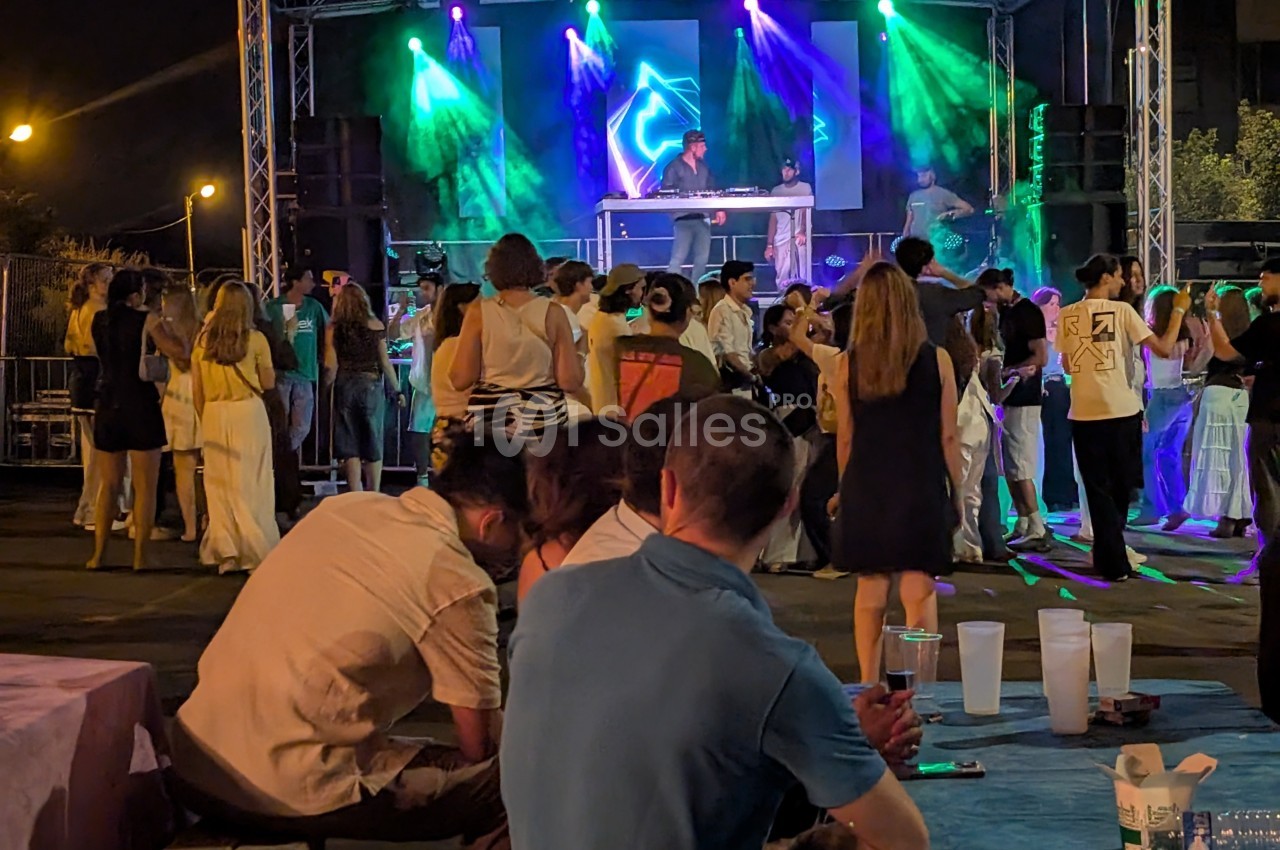 Groupe de personnes dansant et socialisant devant une scène illuminée avec un DJ lors d'un événement en soirée.
