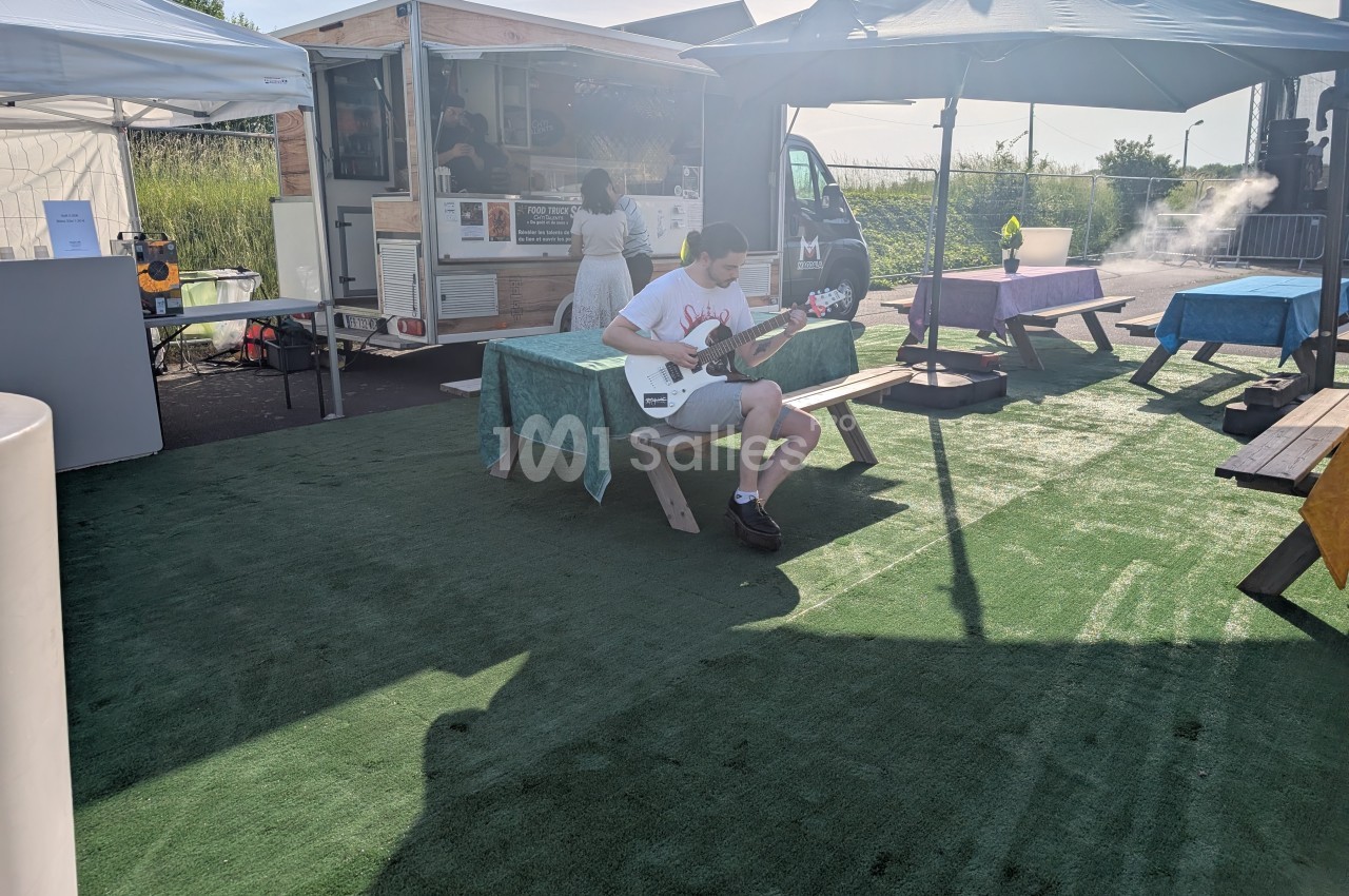 Un homme joue de la guitare assis sur un banc en bois dans un espace extérieur avec food trucks et parasols.