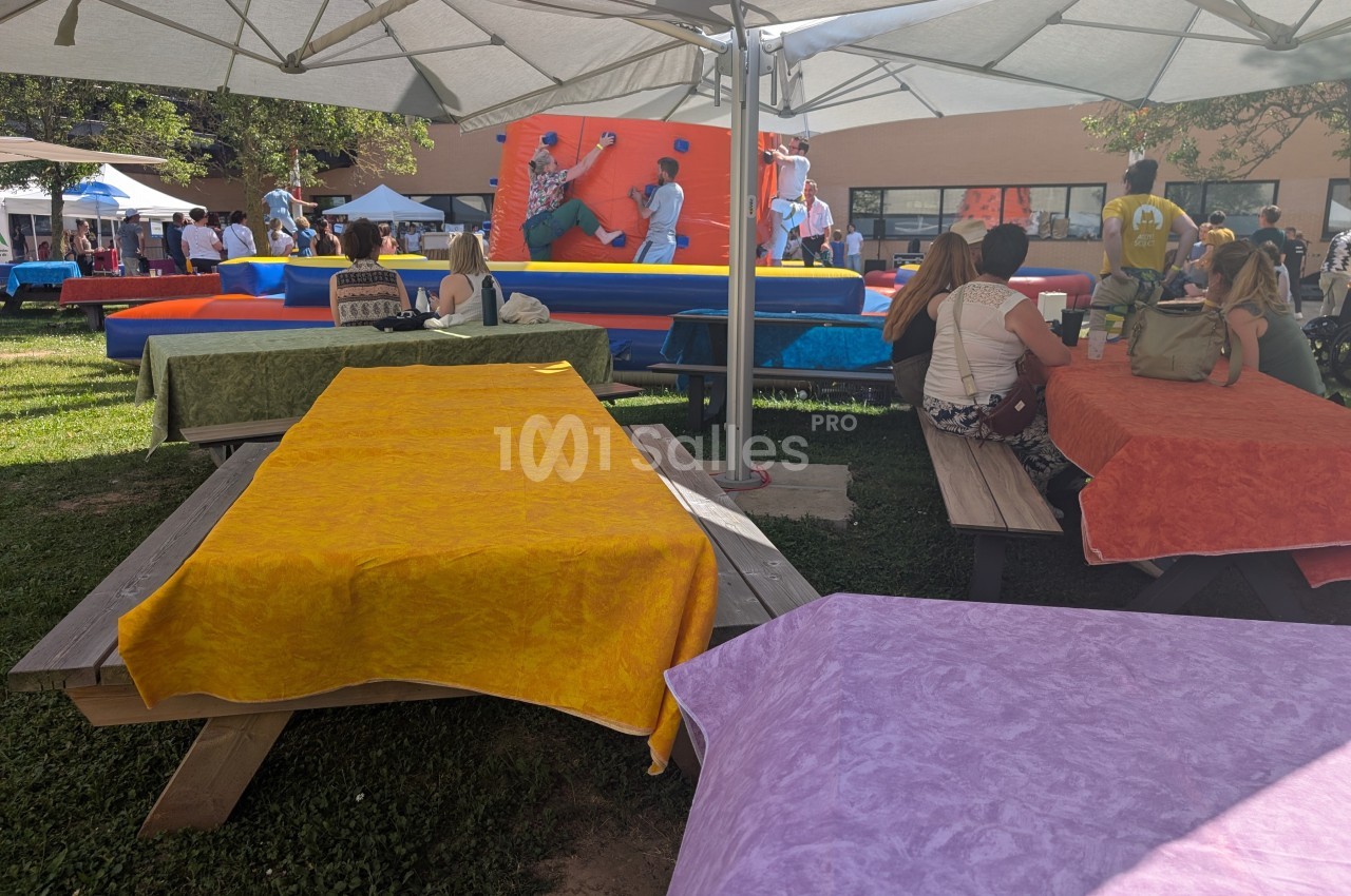 Tables de pique-nique colorées sous des parasols, avec des personnes et une structure gonflable en arrière-plan.