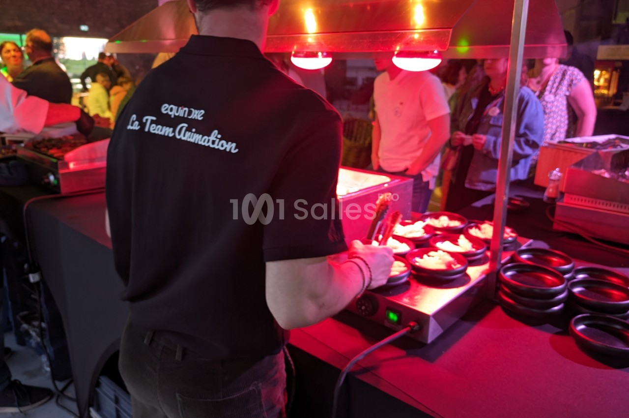 Un cuisinier prépare des plats sous une lampe chauffante lors d'un événement avec des personnes en arrière-plan.