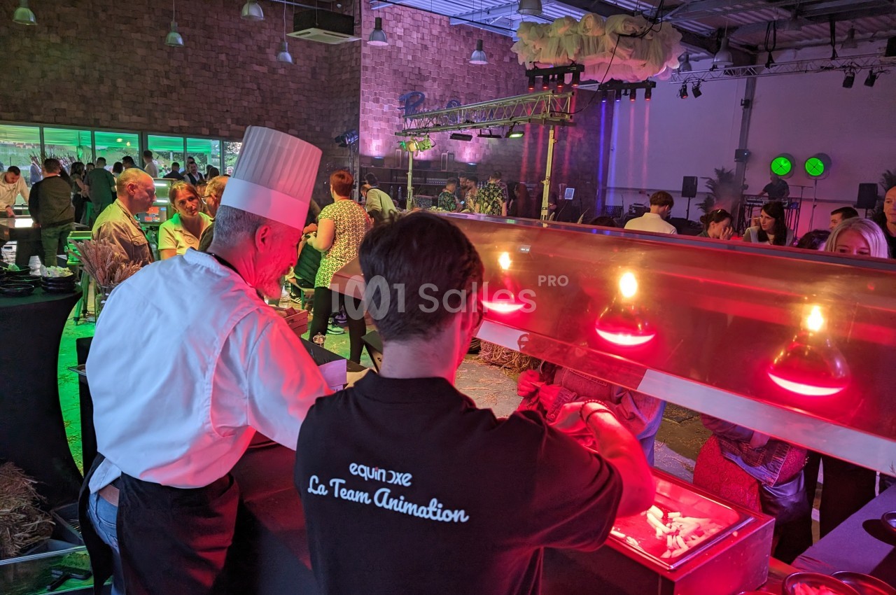 Un chef et un serveur préparent des plats sous des lampes chauffantes lors d'un événement avec des invités en arrière-plan.