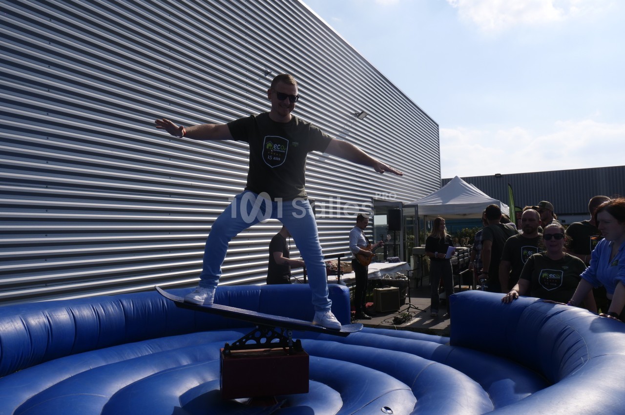 Un homme debout sur un simulateur de surf gonflable lors d'un événement en extérieur, entouré de spectateurs.