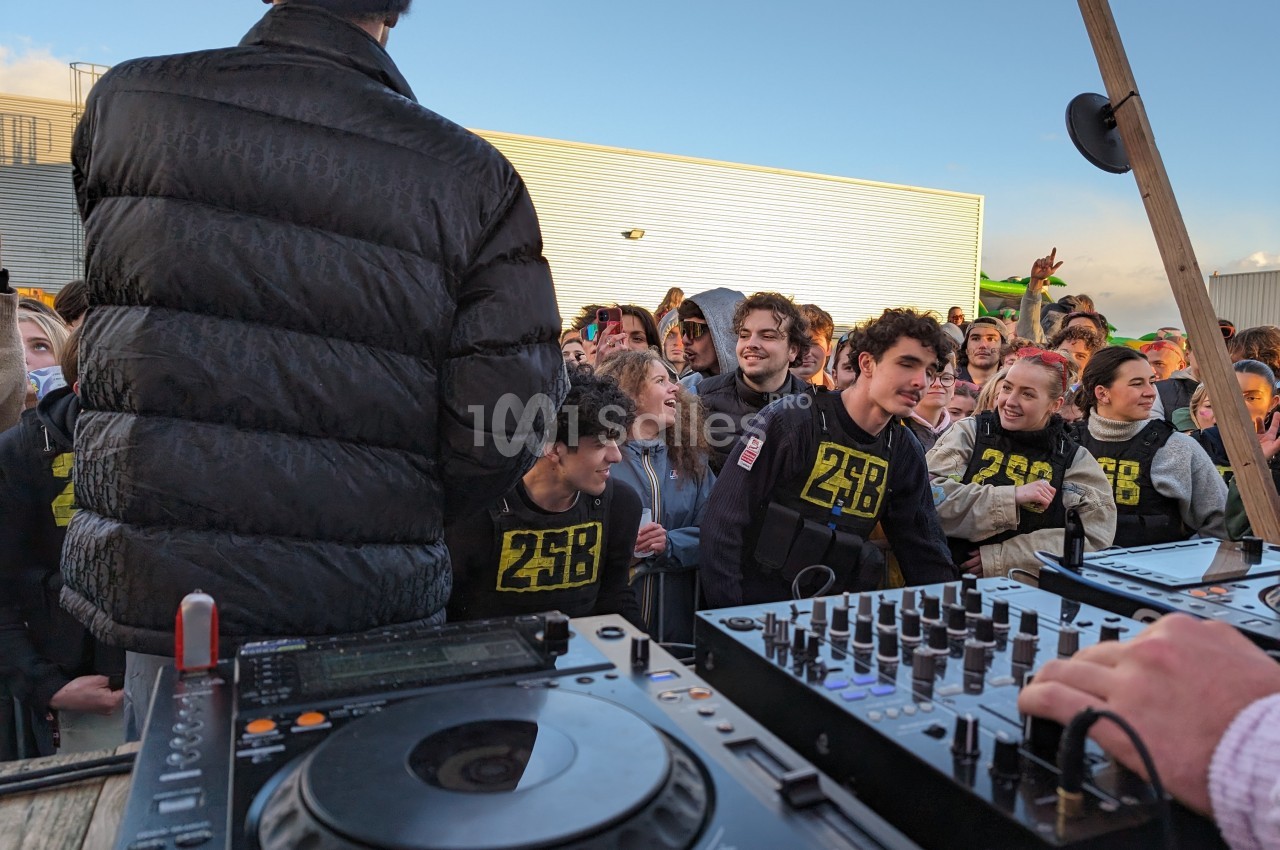 Un DJ anime une foule de jeunes souriants lors d'un événement en extérieur, avec des platines au premier plan.