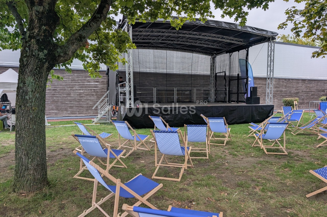 Scène extérieure vide avec des chaises pliantes bleues disposées sur une pelouse sous un arbre.