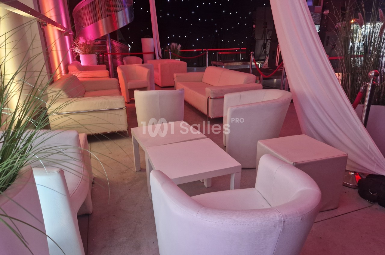 Salon avec fauteuils et canapés blancs disposés autour de petites tables, éclairage tamisé et rideaux blancs.