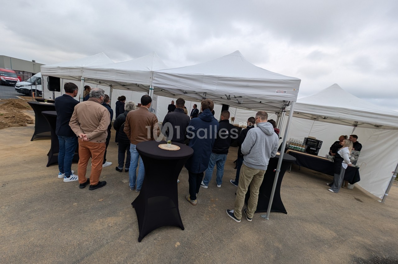 Groupe de personnes debout sous des tentes blanches lors d'un événement en extérieur, avec tables hautes noires.