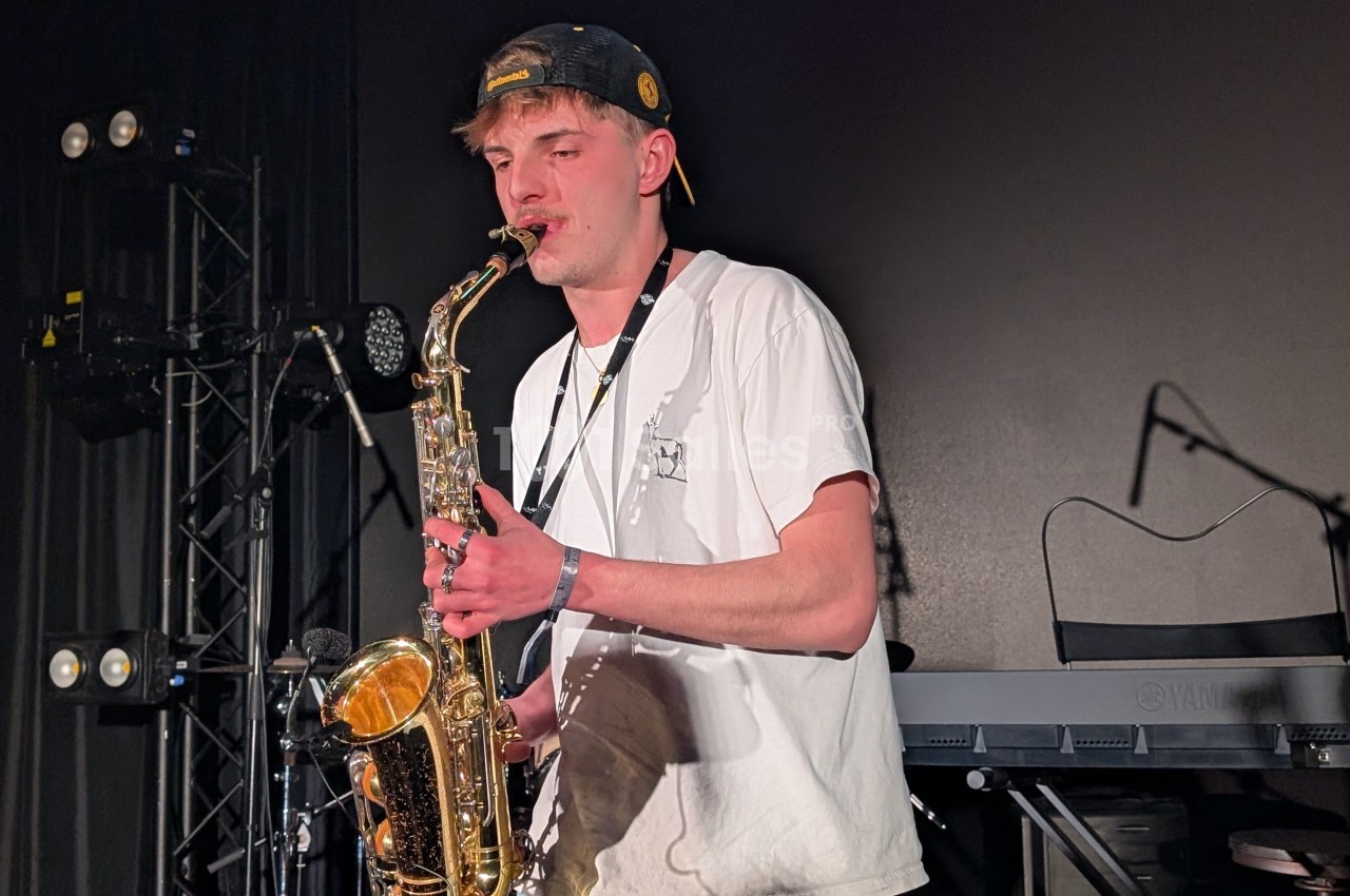 Un musicien joue du saxophone sur une scène sombre, entouré d'équipements de concert.
