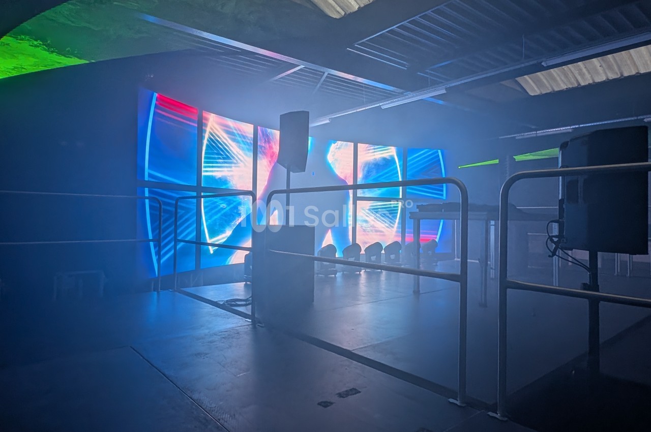 Salle sombre avec écrans lumineux affichant des motifs colorés, entourée de barrières et équipée de haut-parleurs.