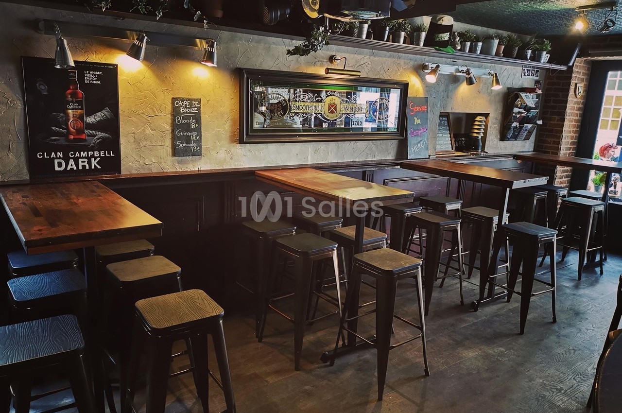 Intérieur d'un bar avec tables hautes en bois, tabourets métalliques et décorations murales dans une ambiance tamisée.