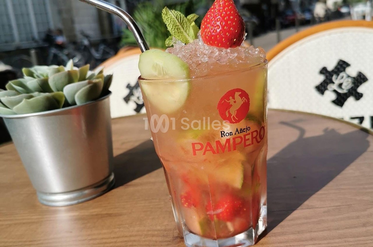 Verre de cocktail fruité avec fraise, concombre, menthe et glaçons, posé sur une table en terrasse ensoleillée.