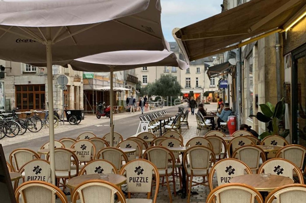 Terrasse de café avec des chaises en osier, tables vides et parasols, située dans une rue pavée bordée de bâtiments.