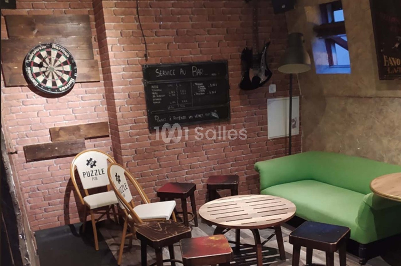 Coin détente avec canapé vert, table basse en bois, chaises, tabourets, cible de fléchettes et mur en briques.