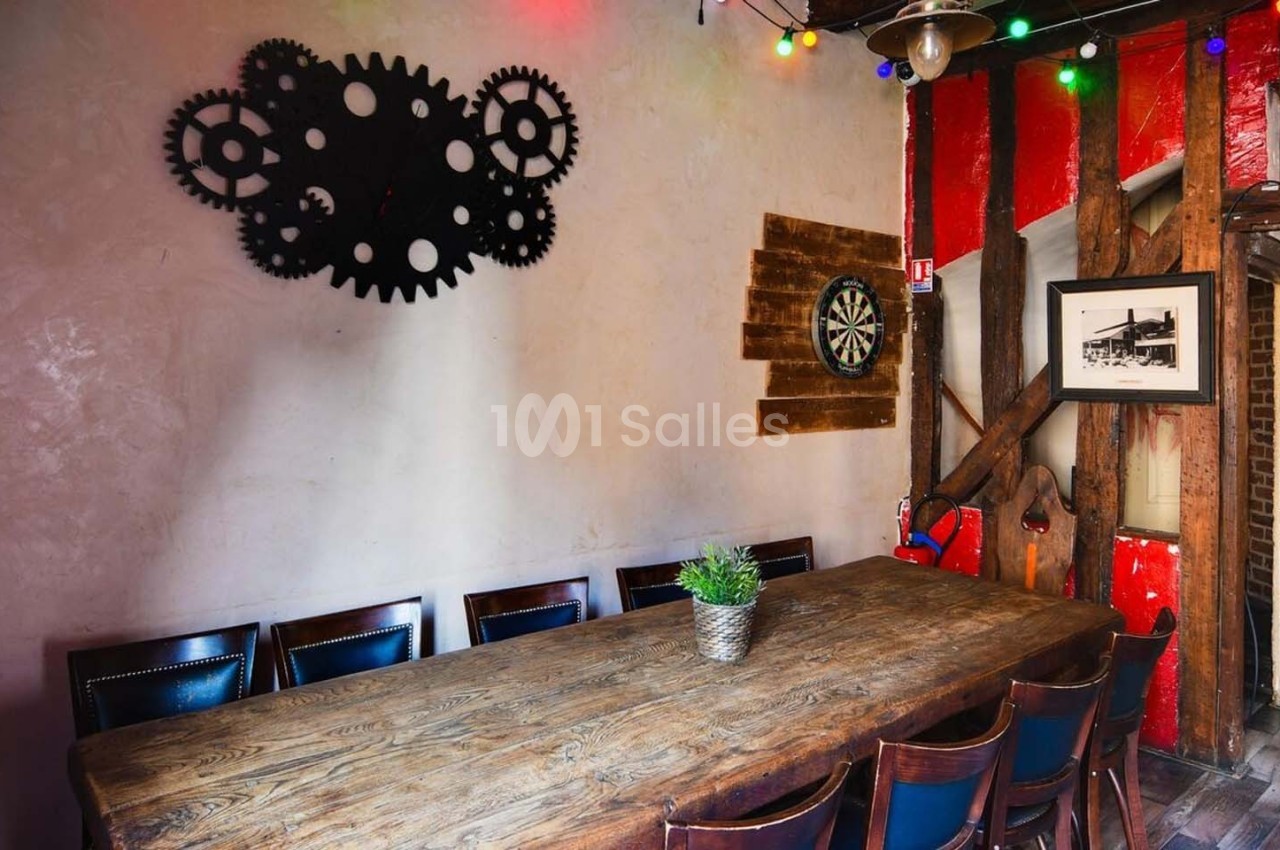 Salle décorée avec une grande table en bois, des chaises, un mur orné de roues dentées et une cible de fléchettes.