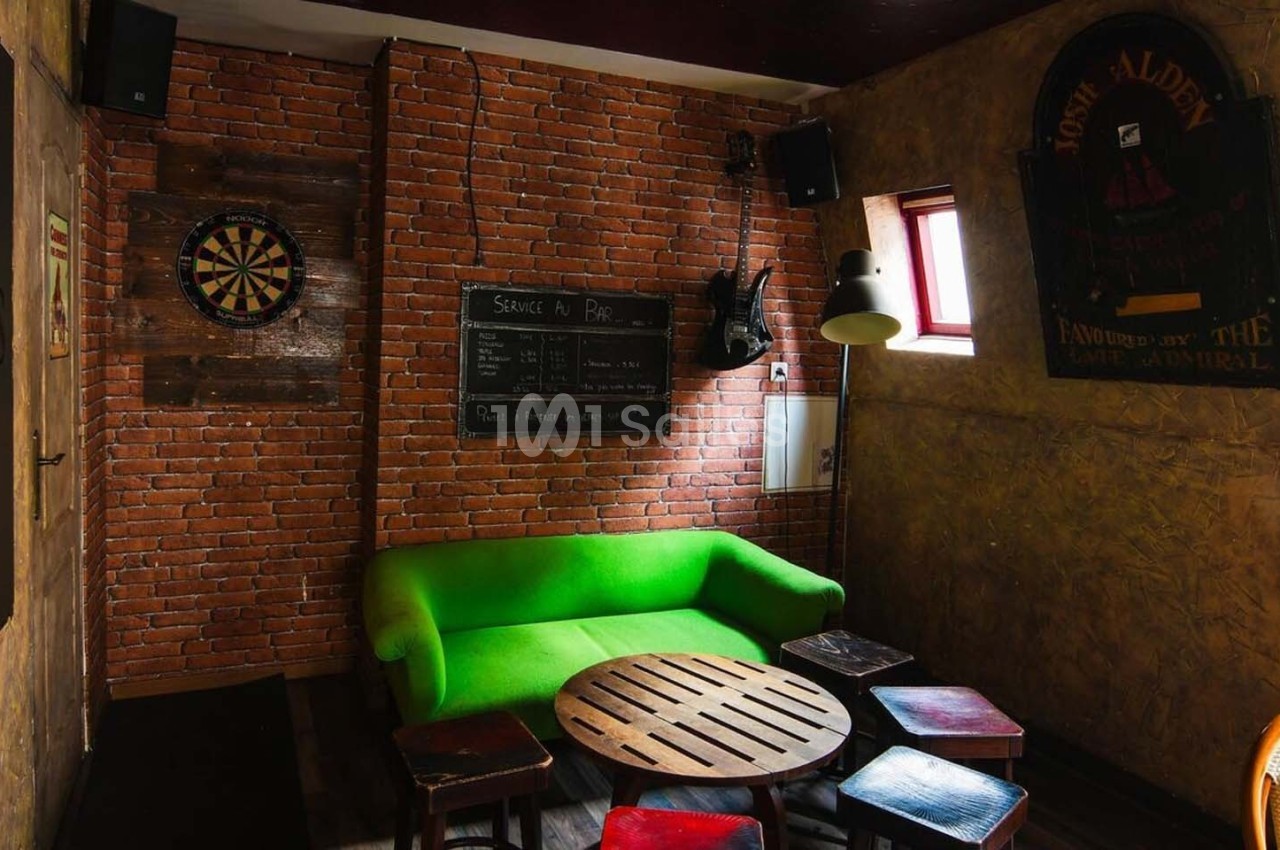 Salle avec murs en briques, canapé vert, table basse en bois, tabourets, cible de fléchettes et tableau noir.