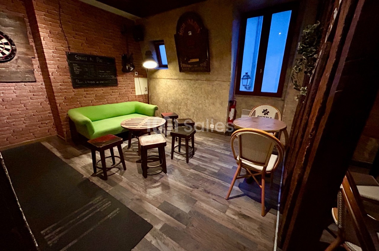 Salle avec murs en briques, canapé vert, chaises et tables en bois, ambiance tamisée et fenêtre donnant sur l'extérieur.