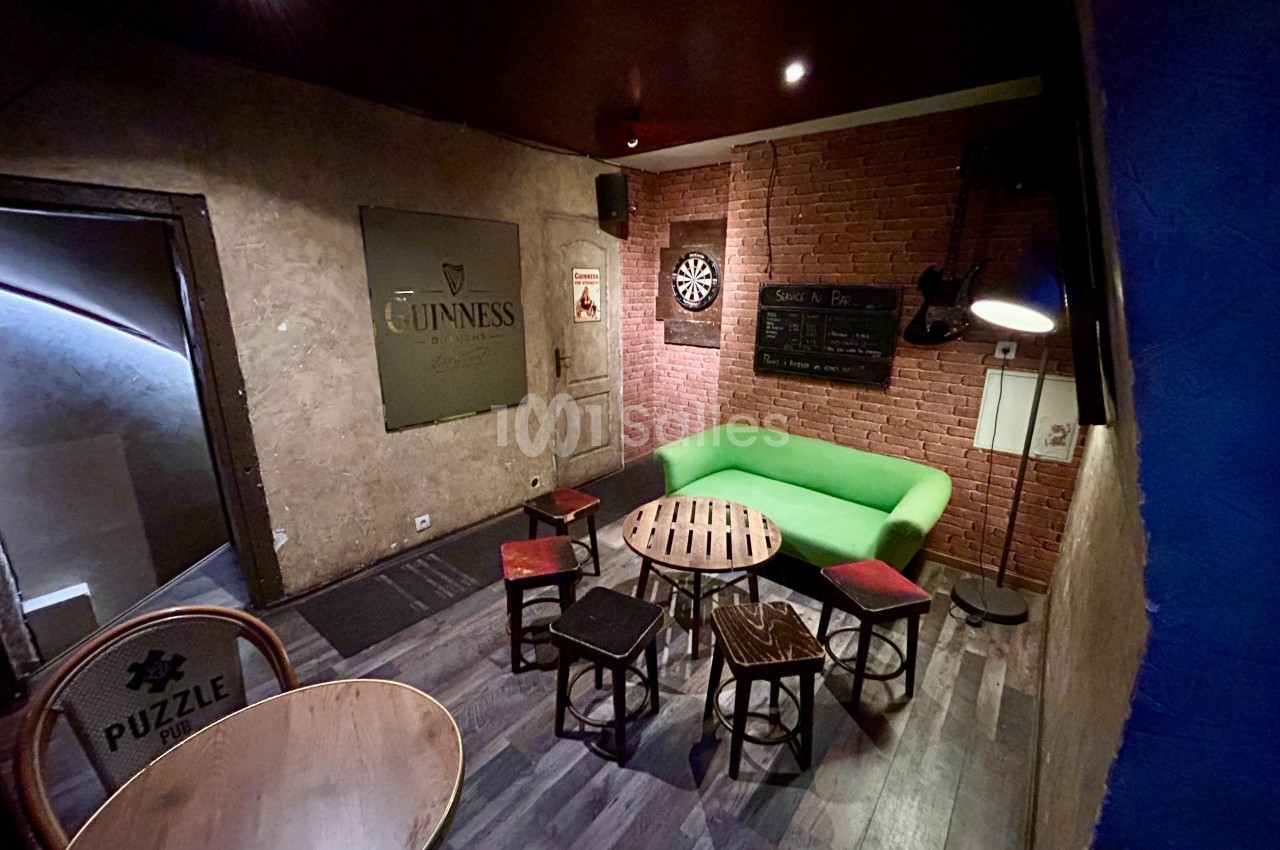 Salle conviviale avec murs en briques, canapé vert, tables en bois, chaises variées et cible de fléchettes au mur.